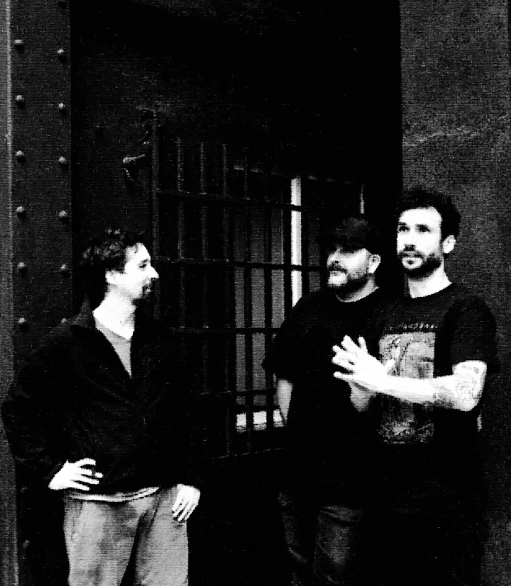 Los tres miembros de REFUGIØ junto a una puerta de hierro con barrotes, en blanco y negro.