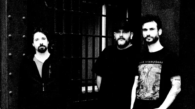 Retrato frontal de los tres miembros de REFUGIØ ante una reja, en blanco y negro.