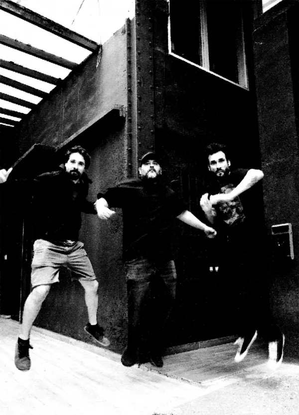 Los tres miembros de REFUGIØ saltando junto a un edificio de acero, fotografiados en contrapicado.